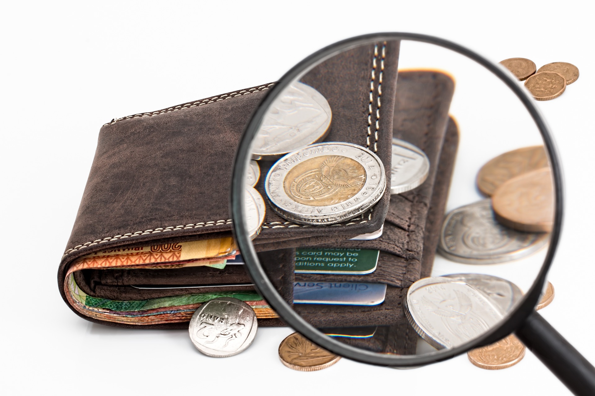 Wallet with coins spread around it with magnifying glass looking at a coin on top of the wallet.